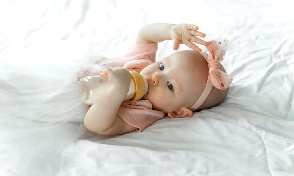 Baby drinks milk from bottle white bed